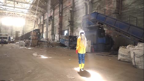 Footage-of-a-woman-with-dreadlocks-wearing-yellow-jacket-and-boots-standing-in-the-middle-of-empty-area-of-waste-recycling-factory-in-rays-of-natural-light-from-cieling