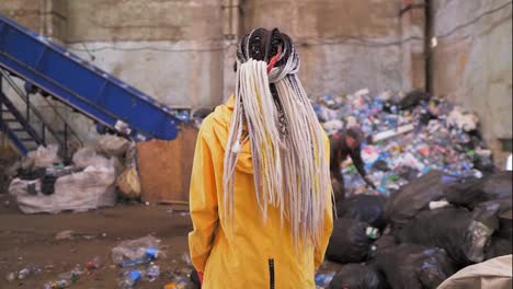 Backside-footage-of-a-woman-with-dreadlocks-wearing-yellow-jacket,-protective-eyeglsses-and-mask-standing-against-waste,-garbage-in-recycling-factory-with-worker-and-equipment-on-background