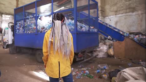 Backside-footage-of-a-woman-with-dreadlocks-wearing-yellow-jacket-watching-the-process-on-waste,-recycling-factory.-Big-truck-with-cage-carcase-of-used-plastic-bottles-for-recycling