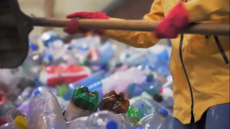 Close-up-footage-of-a-person-in-yellow-jacket-and-gloves-scoops-used-bottles-with-a-large-duck-shovel-at-a-plastic-recycling-factory.-Huge-pile-of-bottles-on-background