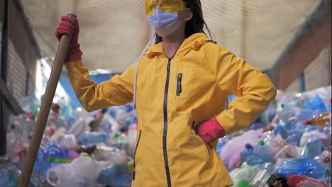 Portrait-of-a-girl-with-dreadlocks,-in-yellow-jacket-and-mask,-holding-large-duck-shovel,-standing-at-a-plastic-recycling-factory.-Huge-pile-of-bottles-on-background.-Low-angle-view,-turning-head-to-the-camera