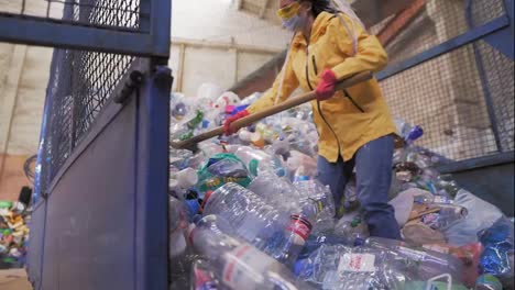 Footage-of-a-young-woman-in-yellow-jacket-and-gloves-scoops-using-shovel-used-plastic-bottles-at-recycling-factory.-Huge-pile-of-bottles-inside-the-car-case-cage.-Slow-motion
