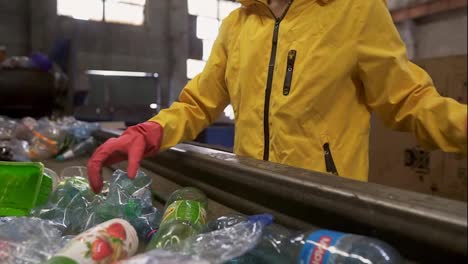 Unrecognizable-person-in-yellow-jacket-and-gloves-sorting-used-plastic-bottles-at-recycling-plant.-Separate-bottles-on-the-line,-squeeze-them-and-put-to-the-box