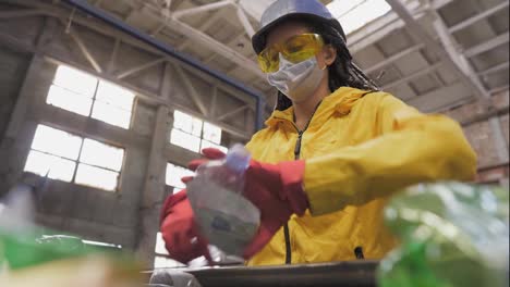 Woman-volunteer-in-yellow-and-transparent-protecting-glasses,-hard-hat-and-mask-sorting-used-plastic-bottles-at-recycling-plant.-Separate-bottles-on-the-line,-removing-tops-and-squeeze-them.-Low-angle-view