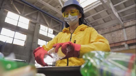 Woman-volunteer-in-yellow-jacket-and-transparent-protecting-glasses,-hard-hat-and-mask-sorting-used-plastic-bottles-at-recycling