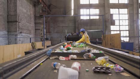 Woman-volunteer-in-yellow-and-transparent-protecting-glasses,-hard-hat-and-mask-sorting-used-plastic-bottles-at-recycling-plant.-Separate-bottles-on-the-line,-removing-tops-and-squeeze-them.Low-angle-footage-from-the-conveyor