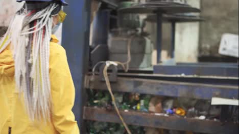 Woman-worker-in-yellow-and-transparent-protecting-glasses,-hard-hat-and-mask-inspect-equipment-on-recycle-plant.-Press-machine-with-used-plastic-bottles-and-differen-plastic-garbage.-Footage-of-automized-process