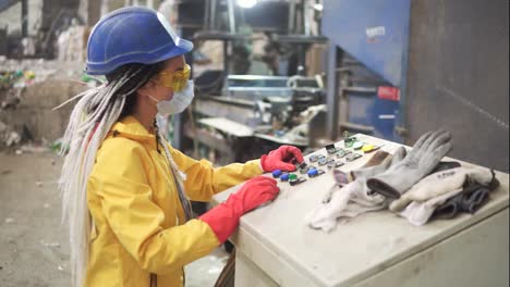 Woman-worker-in-yellow-and-transparent-protecting-glasses,-hard-hat-and-mask-working-with-equipment-on-recycle-plant.-Press-machine-with-used-plastic-bottles-and-differen-plastic-garbage.-Footage-of-automized-process