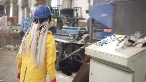 Woman-worker-with-dradlocks-in-yellow-and-transparent-protecting-glasses,-hard-hat-and-mask-working-with-equipment-on-recycle-plant.-Press-machine-with-used-plastic-bottles-and-differen-plastic-garbage.-Footage-of-automized-process