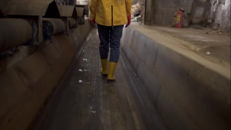 Rare-view-footage-of-a-girl-in-protective-working-clothes---walking-by-moving-conveyor-waste-recycling-factory,-huge-area-with-used-bottles,-different-waste-and-garbage