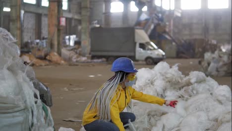 Waste-processing-plant.-Technological-process.-Recycling-and-storage-of-waste-for-further-disposal.-Woman-worker-in-hard-hat-gathering-from-the-floor,-pressing-used-plastic-tape-on-recycle-factory.-Slow-motion
