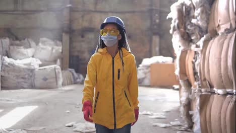 Waste-processing-plant.-Recycling-and-storage-of-waste-for-further-disposal.-Woman-worker-in-hard-hat,-gloves-and-mask-walking-through-the-stacks-of-pressed-disposal-waste,-huge-piles.-Front-view-footage