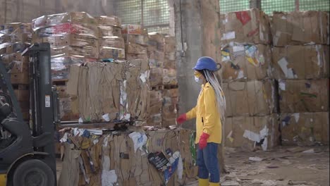 Unloading-of-waste-paper-in-a-warehouse-electric-car-controling-by-female-worker-in-hard-hat-and-yellow-jacket.-Huge-stocks-of-pressed-carton.-Slow-motion