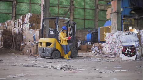 Female-worker-in-hard-hat-and-yellow-jacket-standing,-leaning-on-warehouse-electric-car-and-reading-work-journal.-Huge-stocks-of-pressed-carton.-Full-length