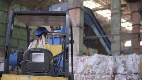 Unloading-of-waste-paper-in-a-warehouse-electric-car.-Female-worker-in-hard-hat-and-yellow-jacket-sitting-in-machine.-Huge-stocks-of-pressed-carton.-Woman-driver-sits-from-the-back-and-pointing-on-smth
