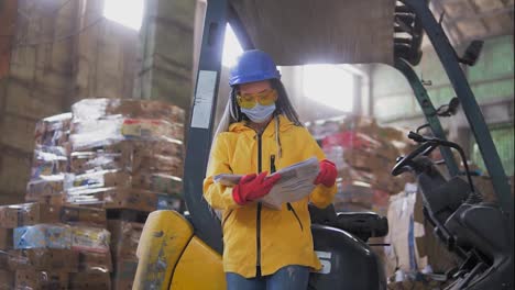 Female-worker-in-hard-hat-and-yellow-jacket-standing,-leaning-on-warehouse-electric-car-and-reading-work-journal.-Huge-stocks-of-pressed-carton.-Low-angle-view