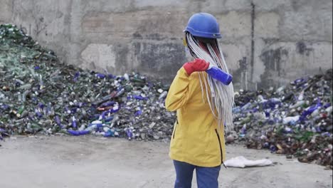 Woman-in-hard-hat-standing-against-the-pile-of-broken-glass,-used-bottles-next-to-the-wall.-Girl-in-yellow-jacket-crashing-old-glass-bottles-for-further-recycling.-Rare-view