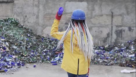 Woman-in-hard-hat-standing-against-the-pile-of-broken-glass,-used-bottles-next-to-the-wall.-Girl-in-yellow-jacket-crashing-old-glass-bottles-for-further-recycling.-Rare-view.-Slow-motion