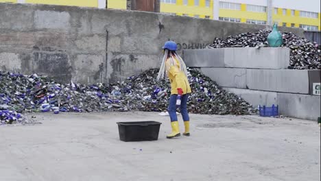 Woman-in-hard-hat-with-dreadlocks-standing-against-the-pile-of-broken-glass,-used-bottles-next-to-the-wall.-Girl-in-yellow-jacket-throwing-crashing-old-glass-bottles-for-further-recycling.-Slow-motion