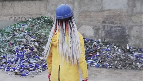Young-woman-in-hard-hat-standing-against-the-pile-of-broken-glass,-used-bottles-next-to-the-wall.-Girl-in-yellow-jacket-crashing-old-glass-bottles-for-further-recycling.-Rare-view.-Slow-motion