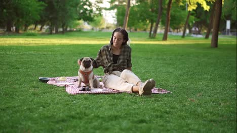 Smiling-girl-sitting-on-plaid-on-lawn-in-a-park-whole-puppy-sitting-in-front-of-her,-she-caress-him