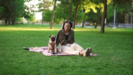 Smiling-girl-sitting-on-plaid-on-lawn-in-a-park.,-the-dog-sitting-in-next-to-her,-she-is-wondering,-looking-at-him