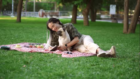 Cheerful-girl-laying-on-plaid-on-lawn-in-a-park-and-making-notes-while-little-pug-sitting-next-to-her