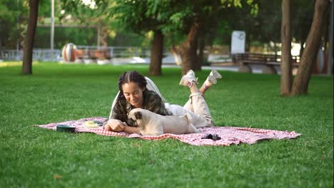 Woman-with-dreadlocks-playing-with-her-dog-on-grass,-laying-on-plaid---tricking-him-with-a-little-snack