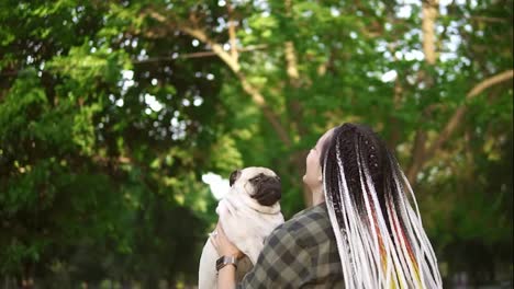 The-girl-and-pug-dog-are-having-fun,-dreadlocks-woman-throws-the-dog-in-the-air-in-the-park