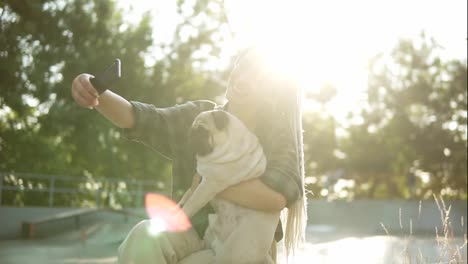 Smiling-girl-taking-selfie-photo-with-cute-pug-puppy-in-green-city-park-holding-smartphone.-Lens-flares