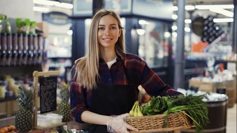 Smiling-supermarket-employee-in-black-apron-holding-a-box-full-of-fruits-and-greens-in-supermarket