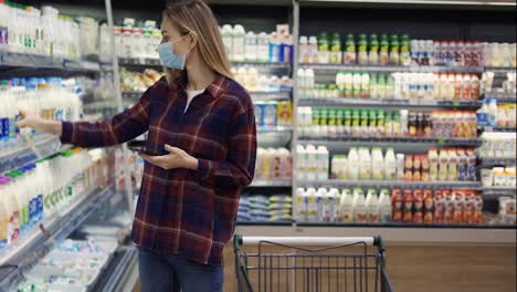Woman-choosing-milk-in-supermarket-using-smart-phone,-checking-shopping-list