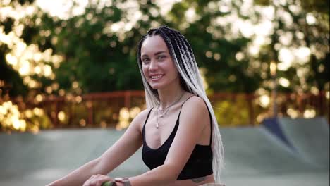 Girl-with-black-and-white-dreadlocks-sitting-on-the-bike-in-skatepark-and-smiling-to-the-camera.-Blurred-trees-in-sunlight-on