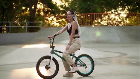 Portrait-of-a-beautiful-girl-with-black-and-white-dreadlocks-sitting-on-the-bike-in-skatepark-and-smiling-to-the-camera