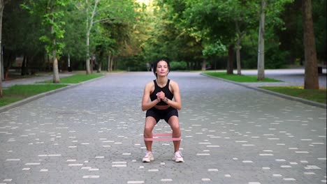 Young-woman-athlete-exercises-with-resistance-band-doing-squats-outdoors-at-local,-green-public-park.-Attractive-woman-wearing