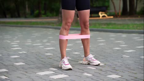 Unrecognizable-young-woman-athlete-exercises-with-pink-resistance-band-doing-squats-outdoors-at-local,-green-public-park