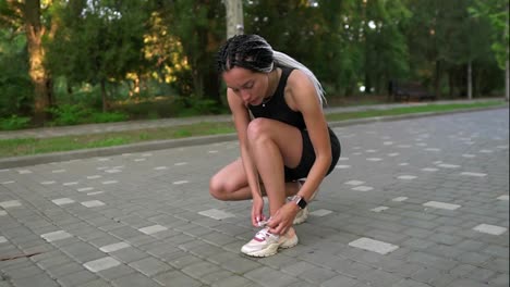 Stylish-woman-runner-with-dreadlocks-in-sport-wear-tying-the-shoelaces-on-pavement-road-in-the-summer-green-park.-Sport-shoes