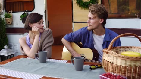 Romantic-couple-sitting-at-the-picnic-table-outdoors-in-front-their-home-trailer-and-man-playing-the-guitar-for-girlfriend.-Girl-listening-music-passionately.-Slow-motion