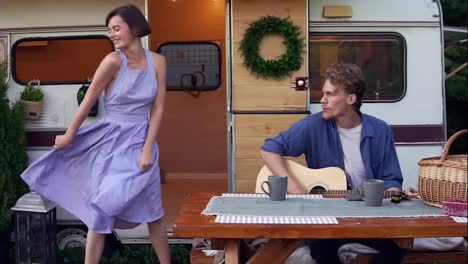 Funny-girl-in-blue-summer-dress-girl-dancing-while-man-playing-guitar-sitting-at-the-wooden-table-in-front-trailer,-singing-a-song-loudly.-Vacation,-holidays,-trailer-trip