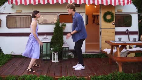 Stylish,-cheerful-couple-man-and-woman-smiling-while-dancing-together-near-house-on-wheels-outdoors.-Funky-dancing-in-front-the-trailer.-Retro-style-exterior.-Slow-motion.-Front-view