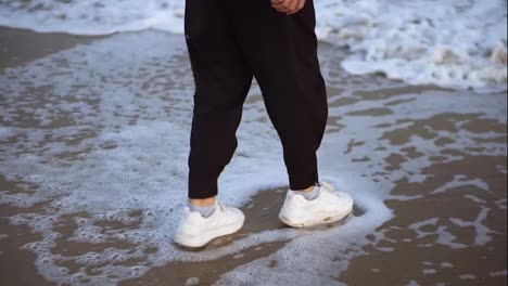 Man's-legs-in-white-sneakers-standing-on-a-sandy-shore-with-waves-coming-at-his-feet