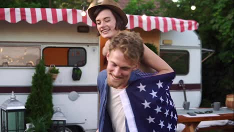 Carefree,-cheerful-couple-having-fun-near-trailer-in-the-park,-man-piggybacking-girl-with-American-flag-on-her-back---spinning-her-around