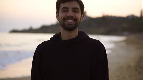 Portrait-of-a-charming-man-smiling-confident-on-calm-seaside-beach-wearing-black-hoodie