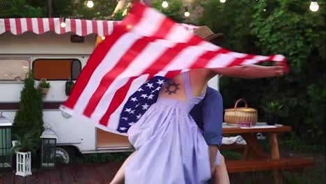 Carefree,-cheerful-couple-having-fun-near-trailer-in-the-park,-man-piggybacking-girl-with-American-flag-on-her-back---spinning-her-around.-Slow-motion