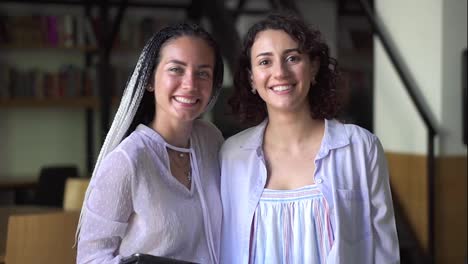 Portrait-of-two-pretty-young-caucasian-women-in-white-blouses-standing-in-library-and-smiling-in-camera,-academic-friends