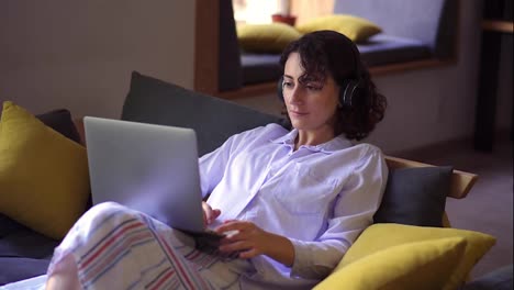 Stylish,-brunette-woman-working-on-laptop-at-home-office.-Young-female-sitting-comfortably-on-a-couch-and-browsing.-Remote-freelance-work-or-studying---in-headphones,-smiling.-Modern-interior