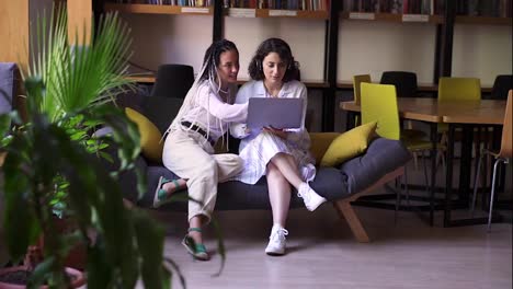 Two-stylish,-focused-female-friends-sitting-on-grey-and-yellow-couch-with-one-laptop,-studying-or-browsing-smth-together.-Two-students-working-remotly-with-one-laptop.-Modern-interior