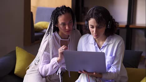 Two-stylish,-focused-female-friends-sitting-on-grey-and-yellow-couch-with-one-laptop,-studying-or-browsing-smth-together.-Two-students-working-remotly-with-one-laptop.-Modern-interior.-Close-up