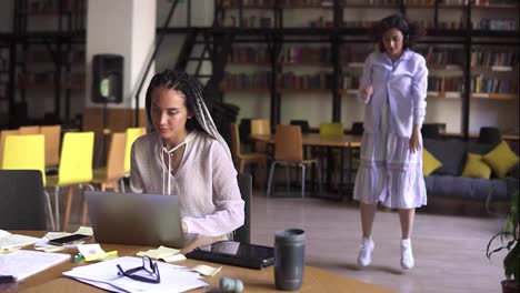 Open-public-library.-Happy-brunette-in-white-casual-clothes-in-headphones-funny-dancing-on-background-while-her-friend-concentrated-on-her-studying,-sitting-at-the-table-works-with-laptop.-Slow-motion
