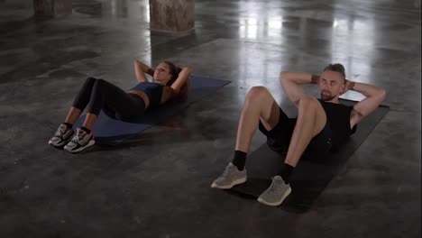 Young-stylish-man-and-woman-in-sportswear-workouting-at-empty-studio,-doing-synchronous-crunches-together,-strong-and-healthy,-maintain-active-lifestyle,-work-hard-to-get-fit,-slow-motion